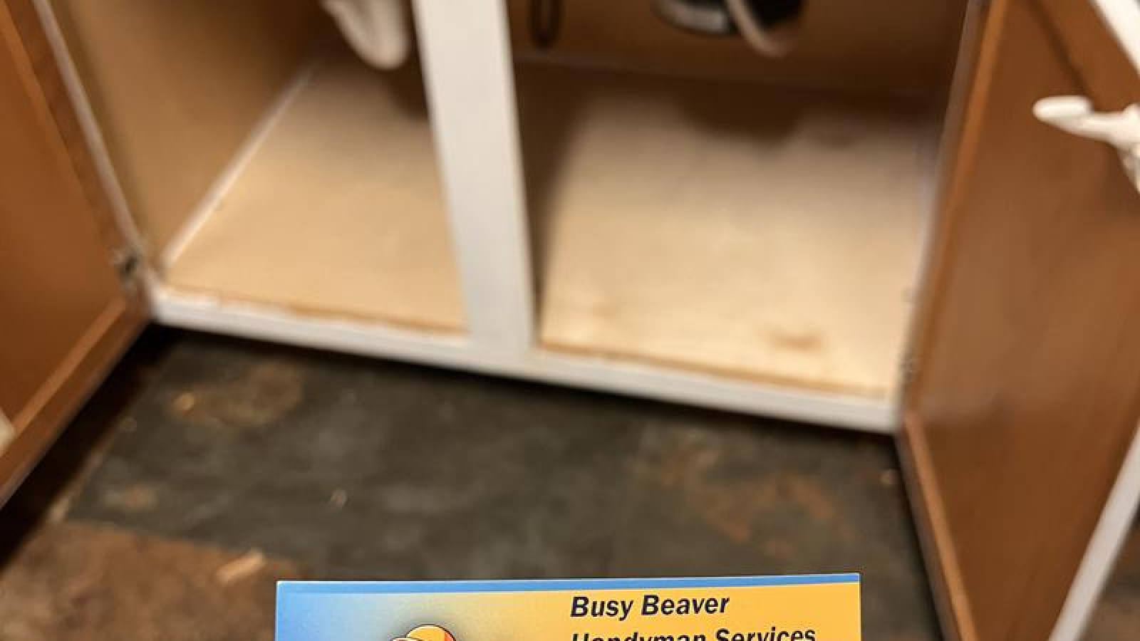 Busy Beaver Handyman Services business card held in front of an open kitchen sink cabinet showing plumbing pipes cabinet repair