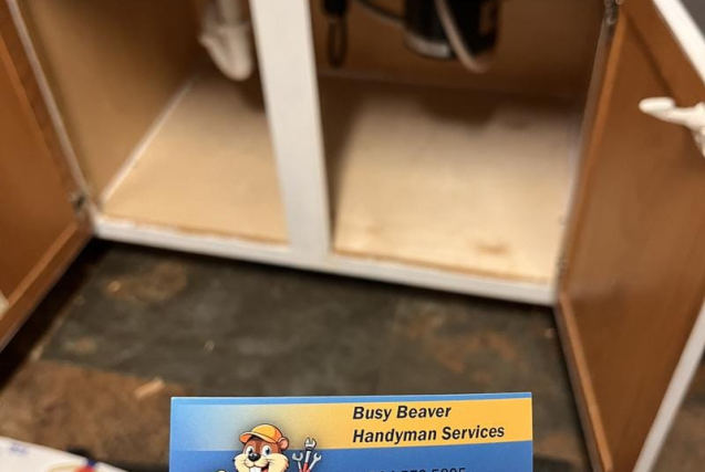 Busy Beaver Handyman Services business card held in front of an open kitchen sink cabinet showing plumbing pipes cabinet repair