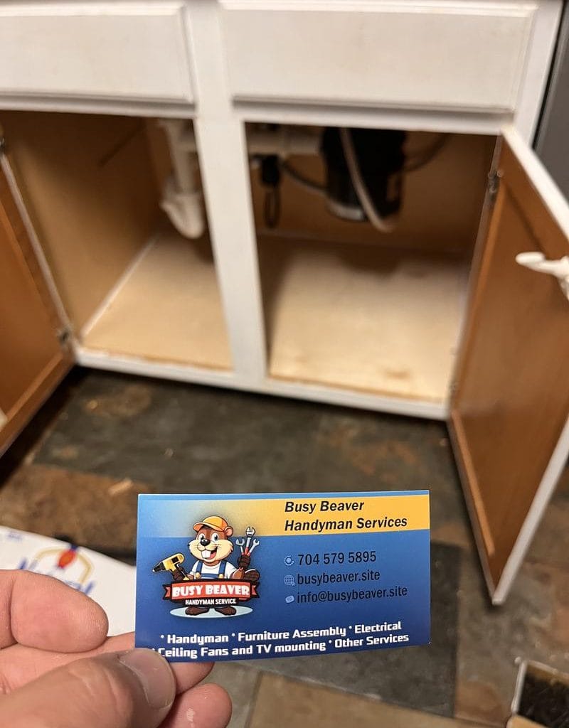 Busy Beaver Handyman Services business card held in front of an open kitchen sink cabinet showing plumbing pipes cabinet repair